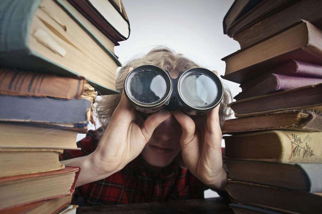 man using binoculars in between stack of books